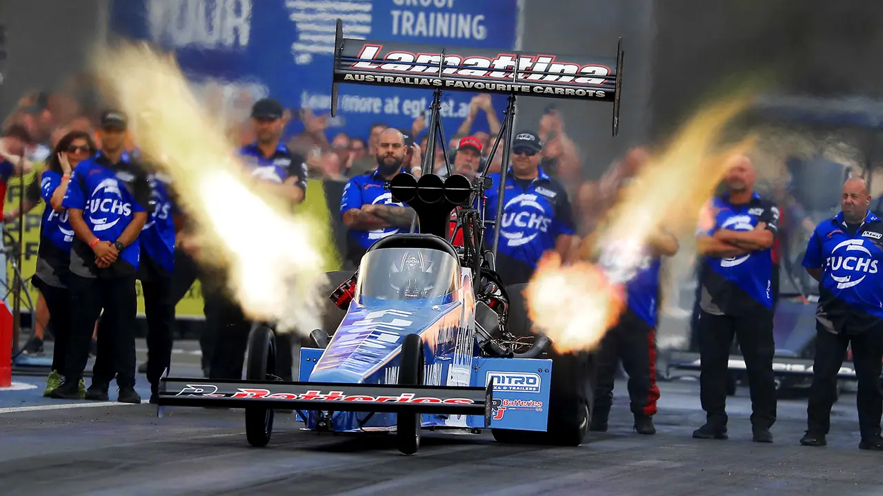 Top Fuel dragster in LTFR and R&J Batteries livery launching with header flames as the FUCHS crew looks on